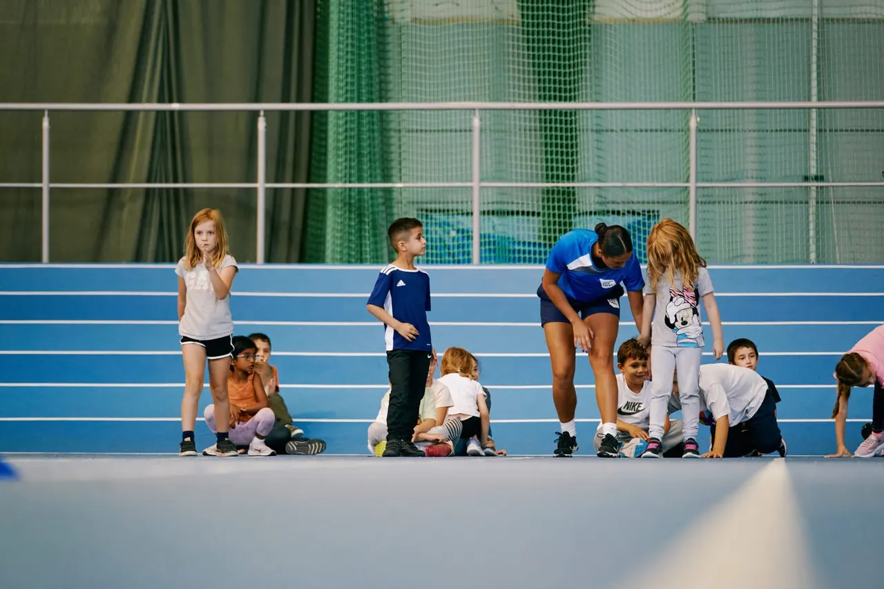 An adult instructor leads a group of young children in a physical activity session on an indoor athletic track.
