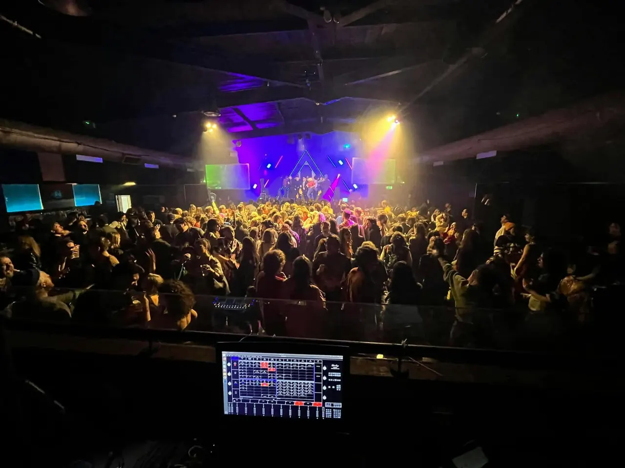 Large crowd dancing and enjoying live music on a stage lit with colourful purple, blue and yellow lights in an indoor venue.