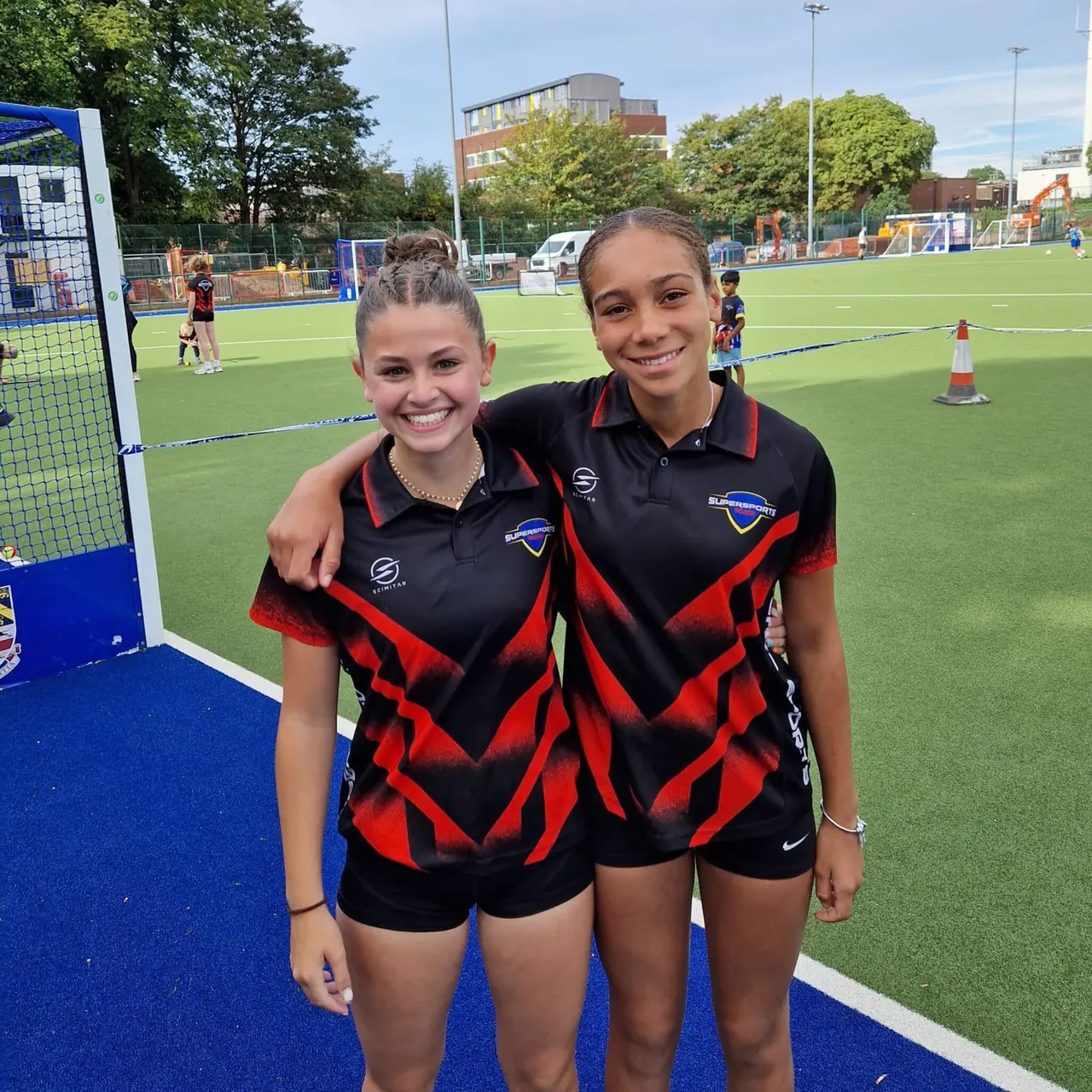 Two young female hockey players in red and black team uniforms smile at the camera on an outdoor astroturf pitch with other players training in the background.