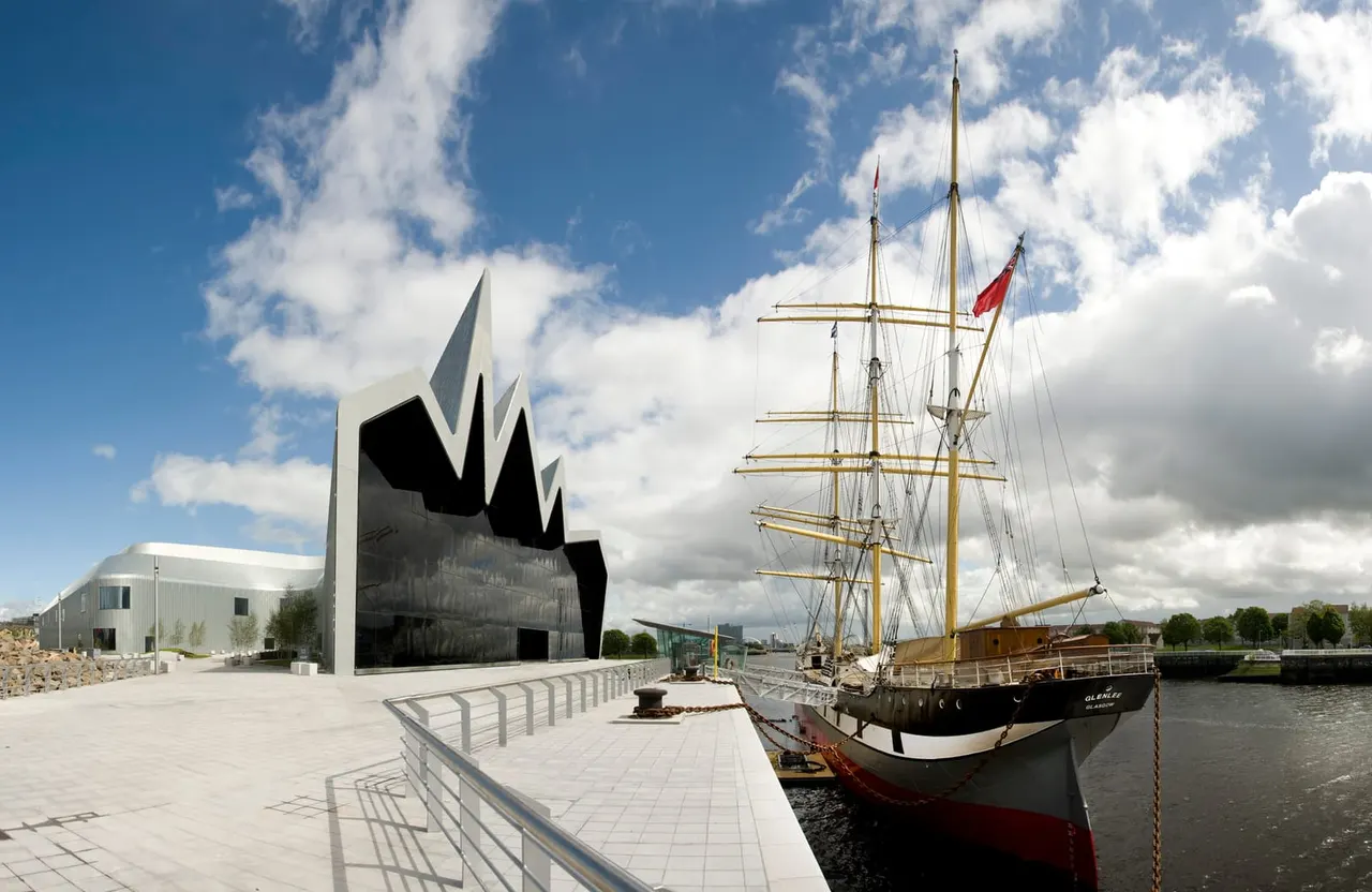 A tall ship with multiple masts moored alongside a modern waterfront building with distinctive angular architecture.