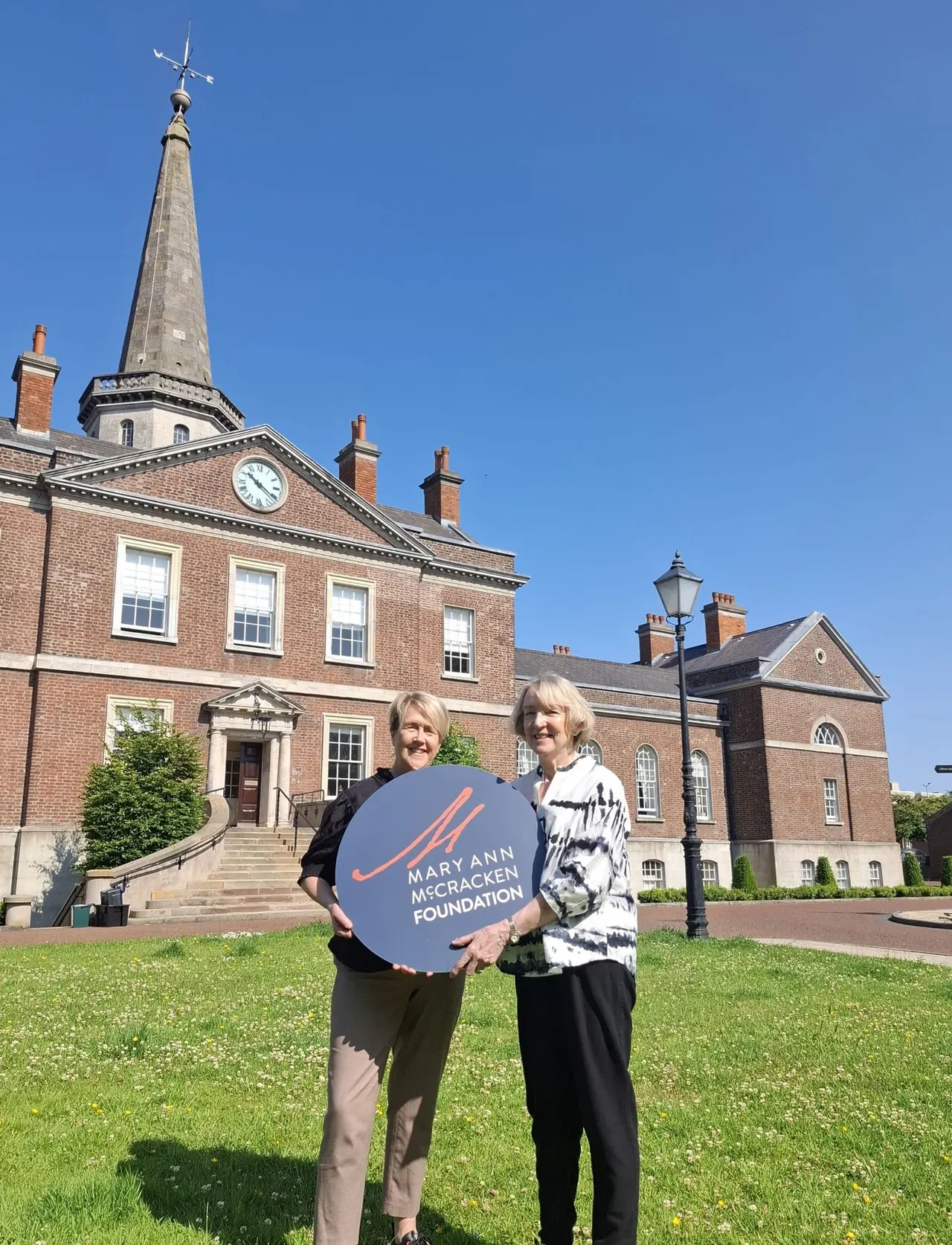 Two women hold a Maryann Mccracken Foundation sign on the lawn in front of a brick Georgian building with a church spire.