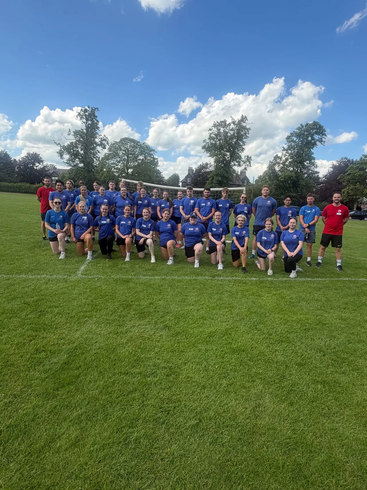 Large group of people in blue and red sports shirts posing on a grass field with volleyball net and trees in background.