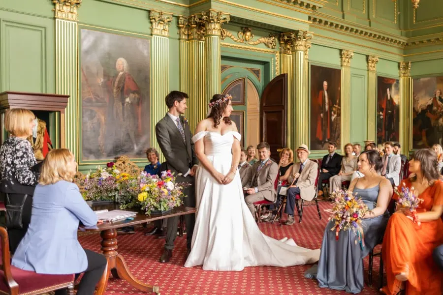 A bride and groom exchange vows during a wedding ceremony in an ornate green-walled hall with gilt columns, surrounded by seated guests.
