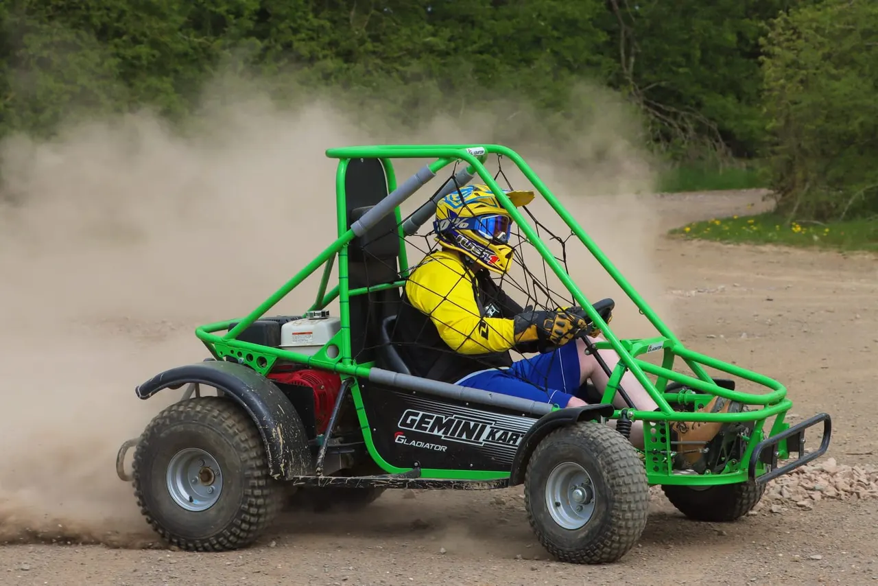 A person in a yellow helmet and blue clothing driving a bright green buggy across dusty terrain with trees in the background.