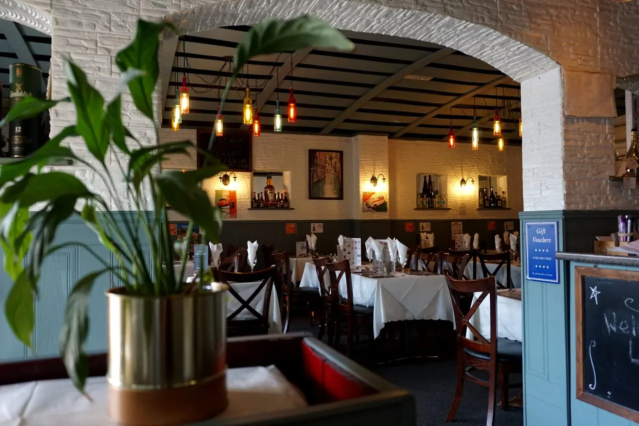 Restaurant dining room with white tablecloths, wooden chairs, hanging bottle lights, brick walls, and potted plant in foreground.