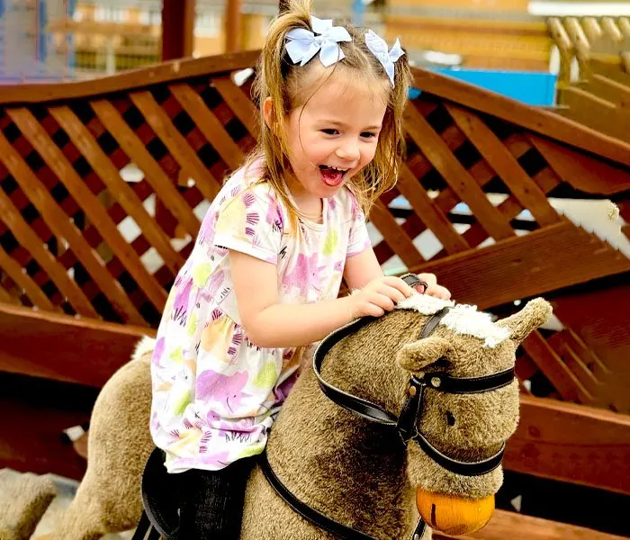 Young girl sitting on a mechanical camel ride, smiling with white hair bows.