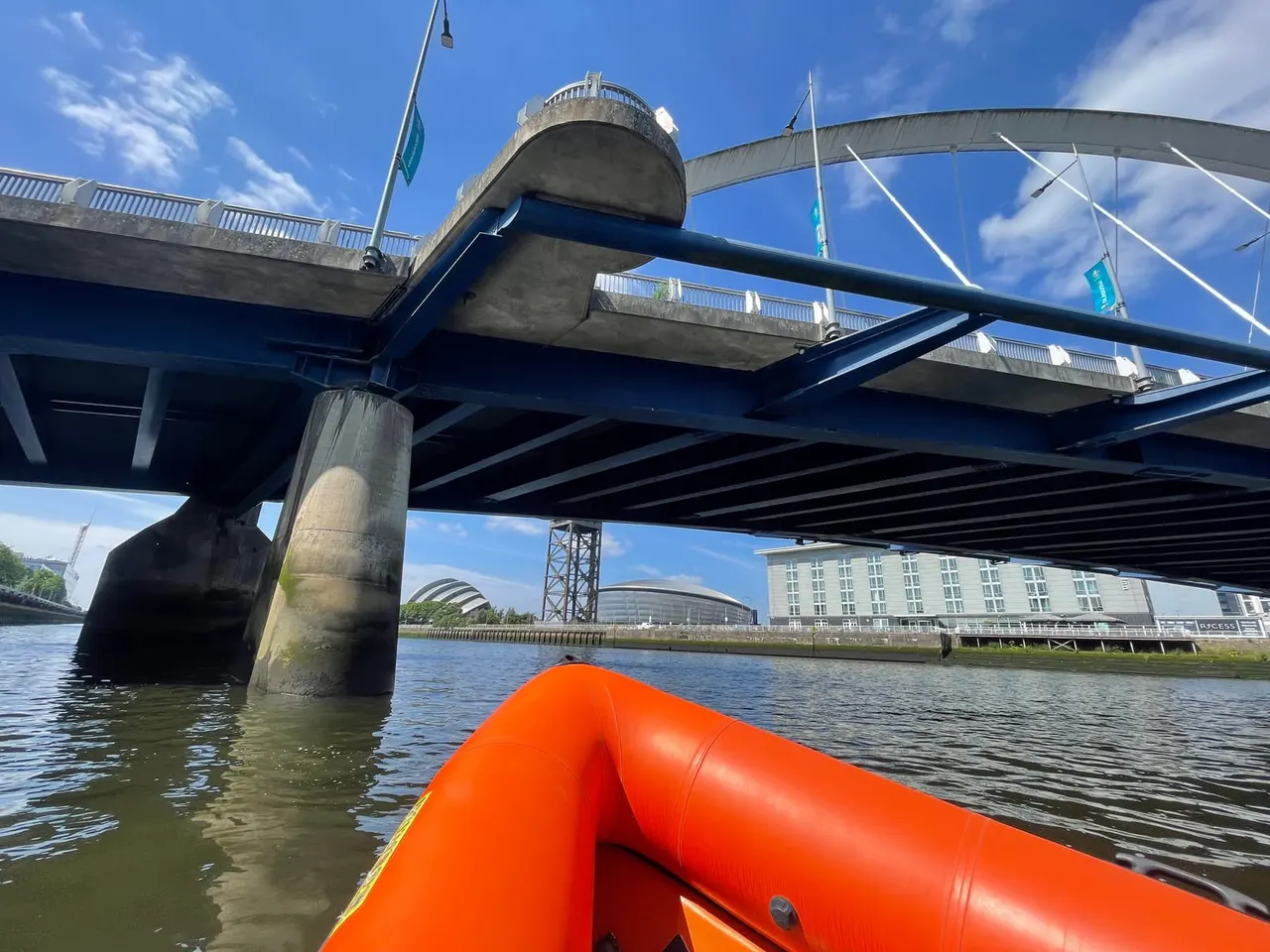 View from an orange inflatable boat passing under a modern pedestrian and cycle bridge spanning a river.