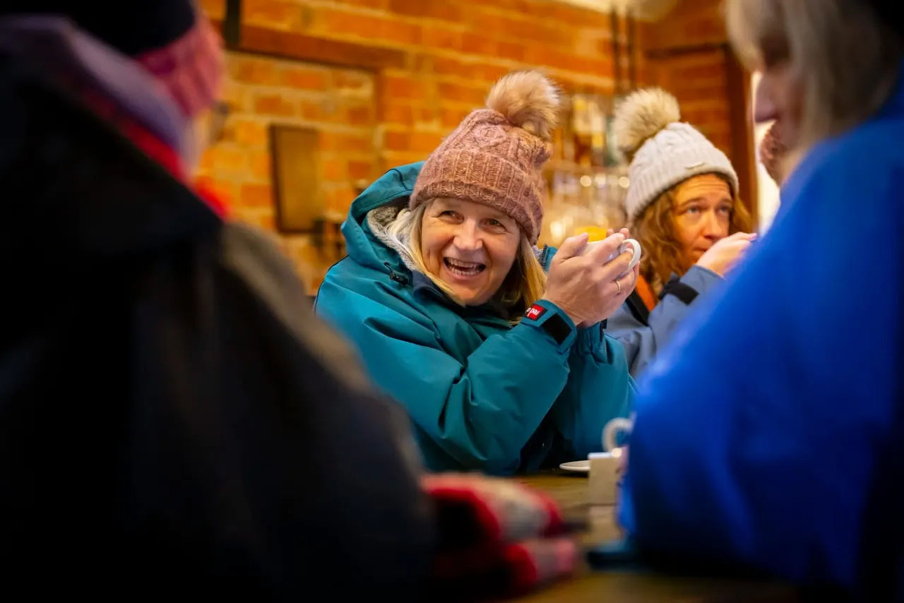 A smiling woman in a teal jacket and pink knitted hat with a pompom sits indoors surrounded by other visitors wearing winter clothing, inside a wooden-beamed building.