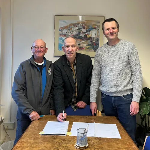 Three men standing at a wooden table with documents, smiling at the camera indoors.