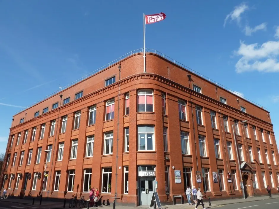 Victorian red-brick corner building flying a flag, with people on the street outside the ground-floor entrance on a clear day.