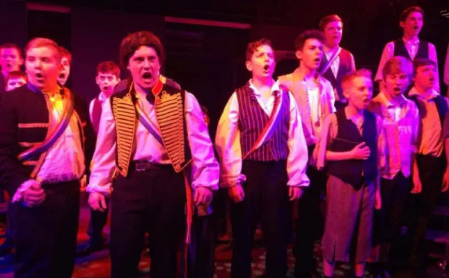 Large group of young performers singing on stage under pink and purple stage lighting, wearing coordinated waistcoats and formal attire.
