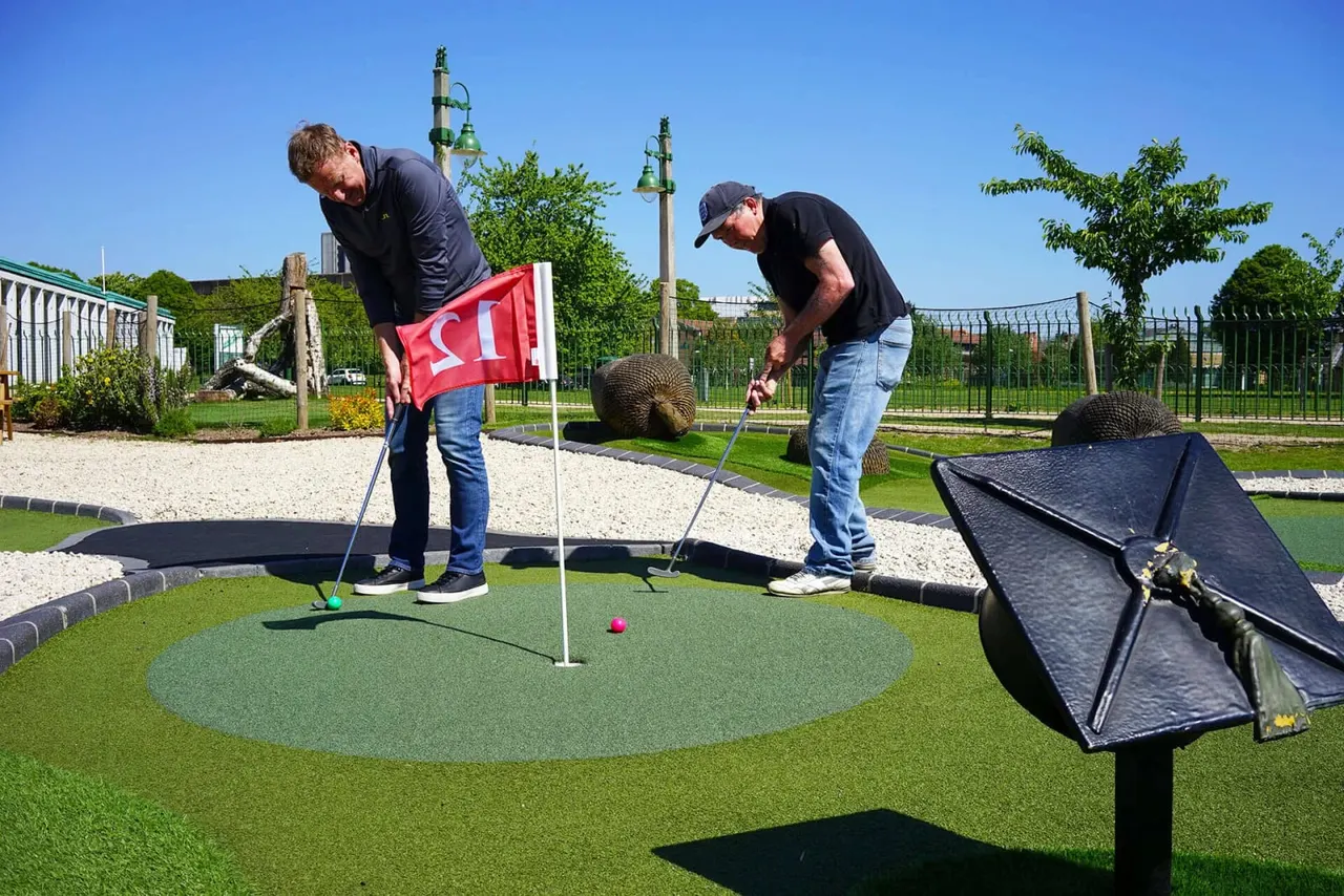 Two men playing mini golf on a bright sunny day at an outdoor course with flags and obstacles.