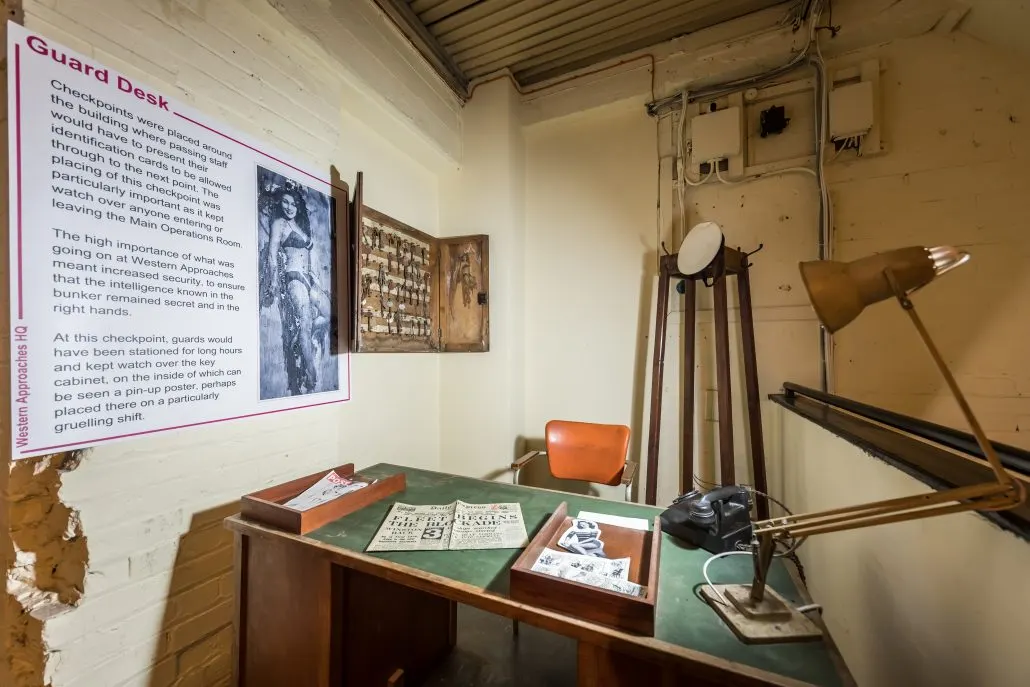 Interior of a historic guard desk with informational panel, vintage desk, chair, lamp and period documents on display.