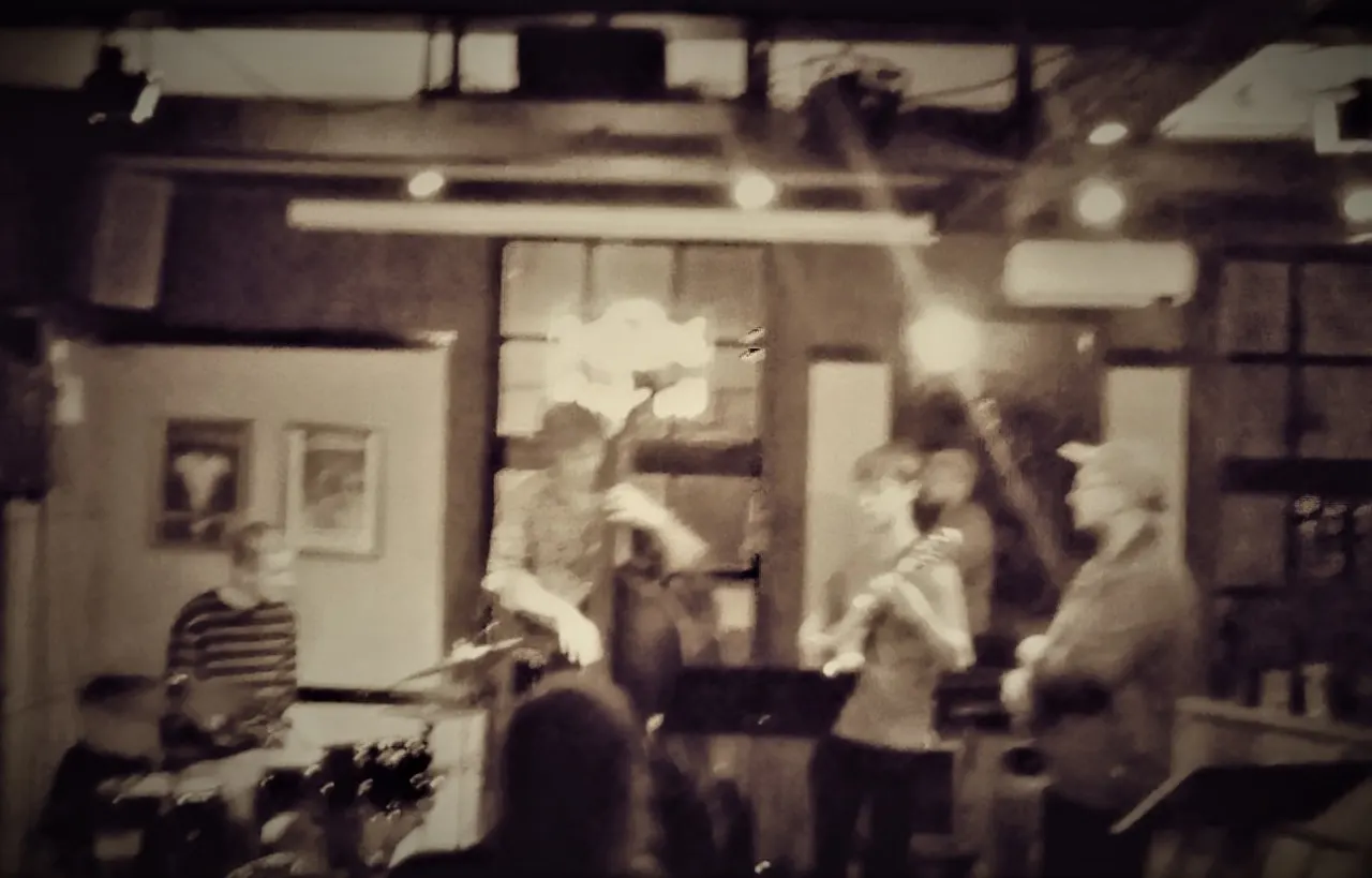 Grainy sepia-toned photograph of people gathered inside a pub or bar with period furnishings and lighting.