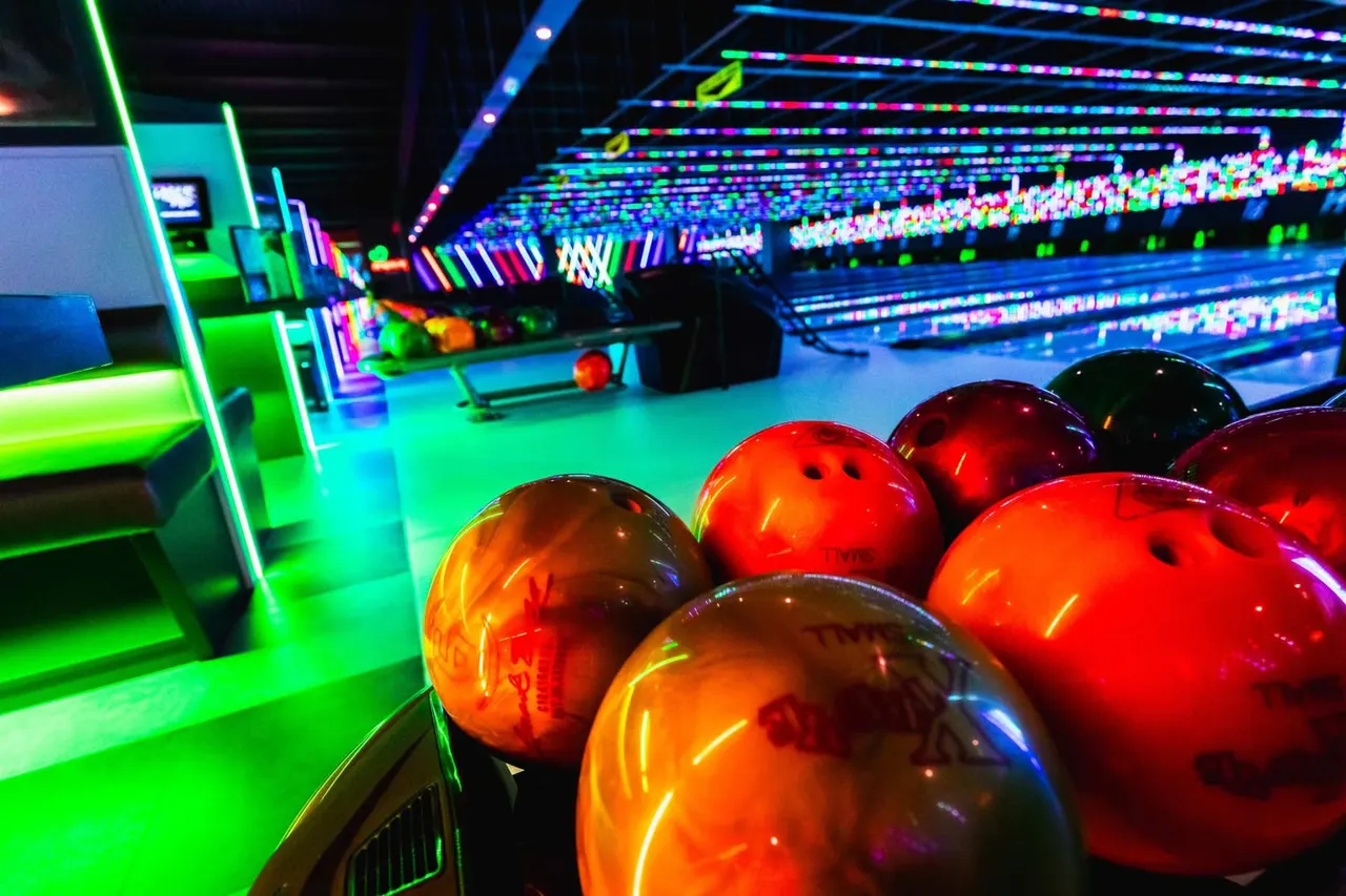 Bowling balls in vibrant red and orange sit on the lane of a neon-lit bowling alley with glowing pins and colourful overhead lighting.