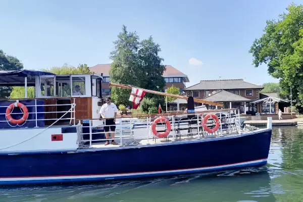 A blue and white passenger riverboat moored on the Thames with crew members on deck and countryside buildings visible along the bank.