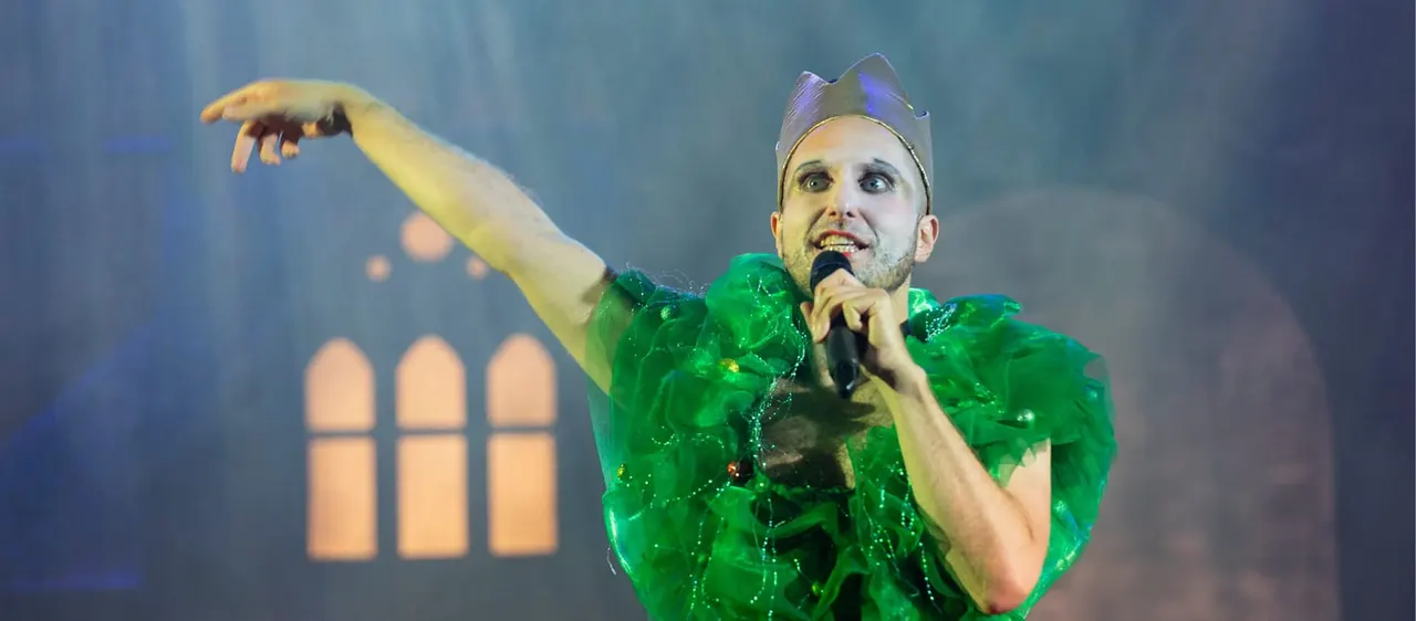 Performer in elaborate green sequined costume and crown singing into microphone on stage with theatrical backdrop.