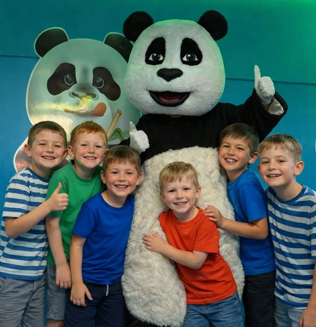 Group of young children posing with a panda mascot character at an indoor play centre.