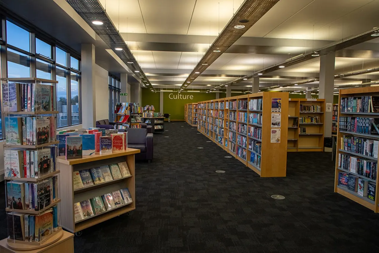 Library culture section with wooden bookshelves, display tables, and seating area beneath large windows.