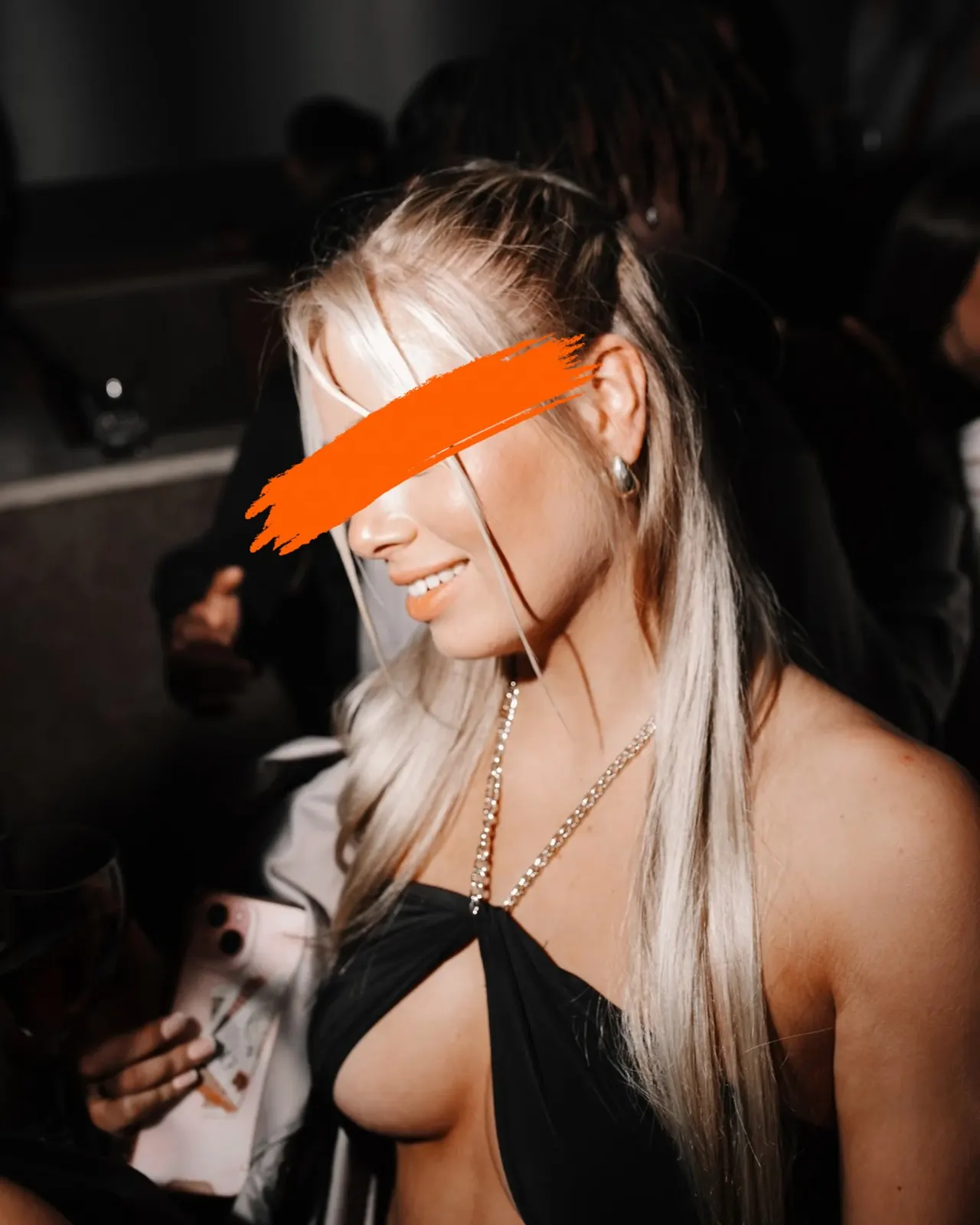 Woman wearing an orange eye mask at an indoor evening event with other guests in the background.