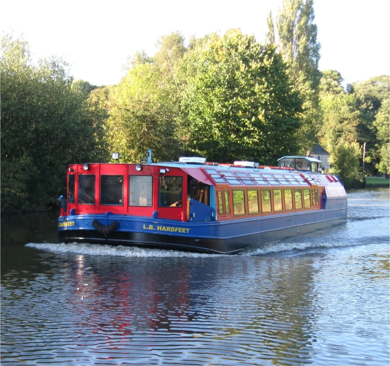 A red and blue passenger riverboat named L.B. Hardfeet on a river with trees lining the banks.