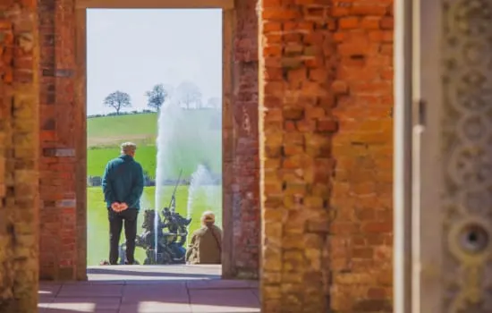 Two people stand outside a brick building doorway, viewing a large garden fountain with green fields and trees beyond.