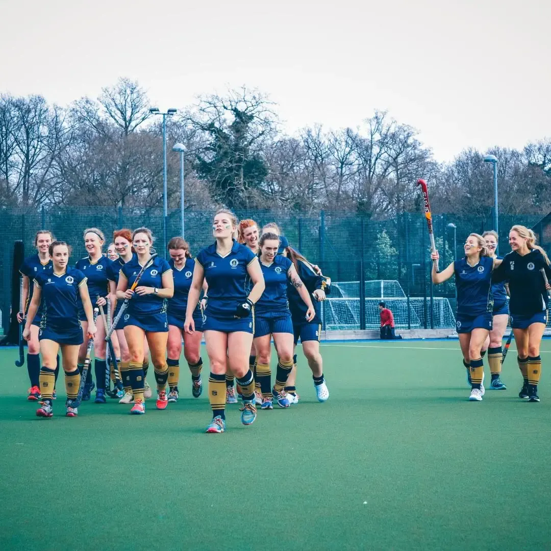 Female field hockey team in blue and gold kit jogging together on an outdoor synthetic pitch during winter.