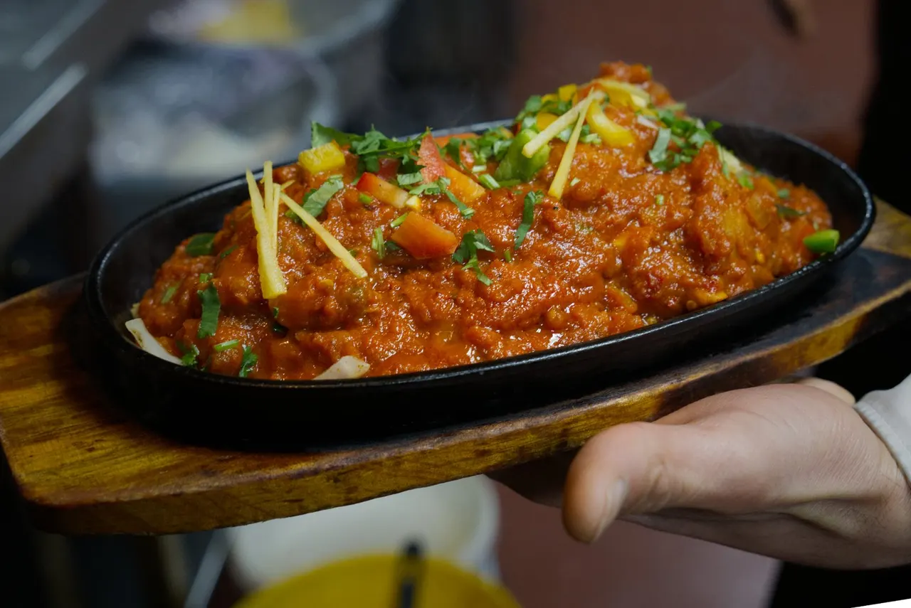 A server holding a sizzling cast-iron skillet of spiced meat curry garnished with ginger and coriander.