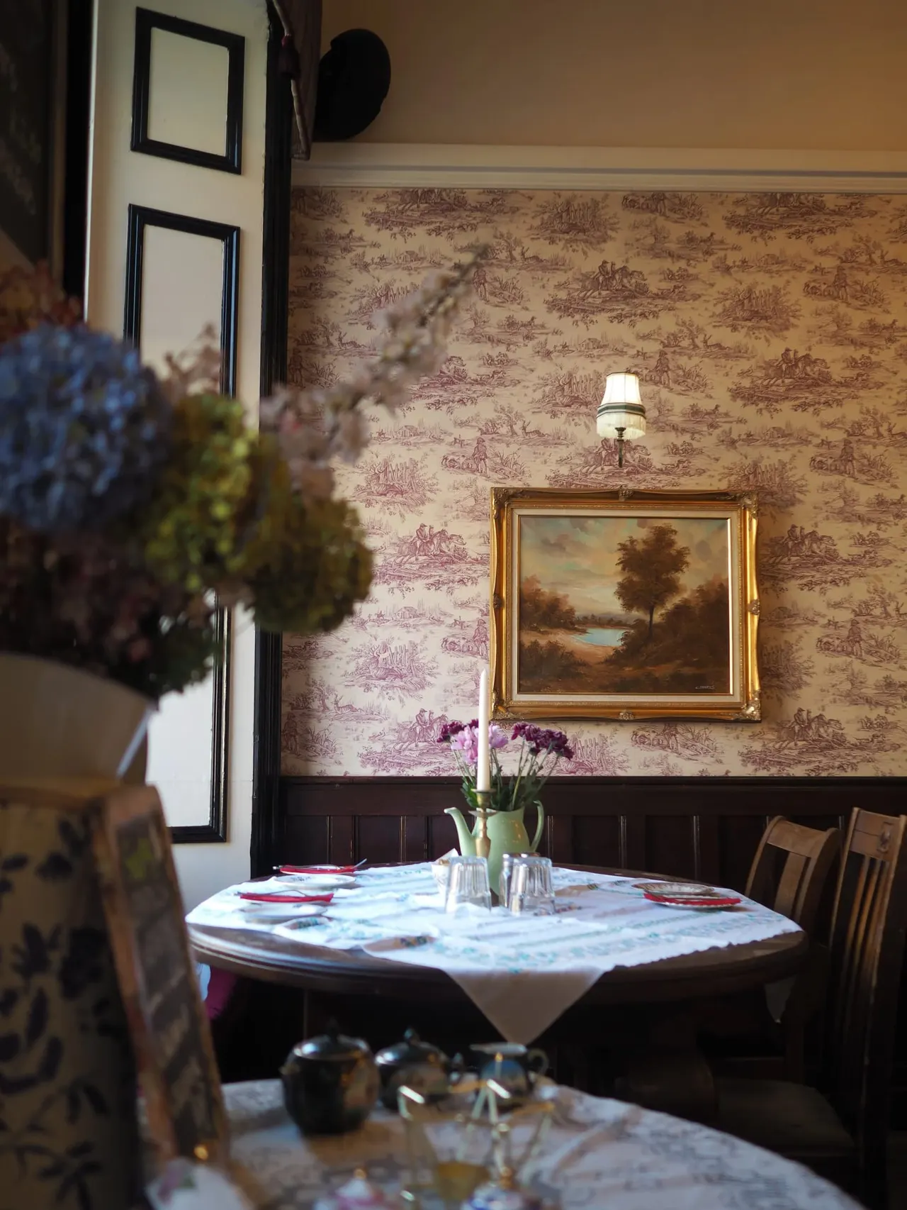 A traditional dining room with a round table set for a meal, featuring toile wallpaper, framed landscape artwork, and fresh flowers in a vase.