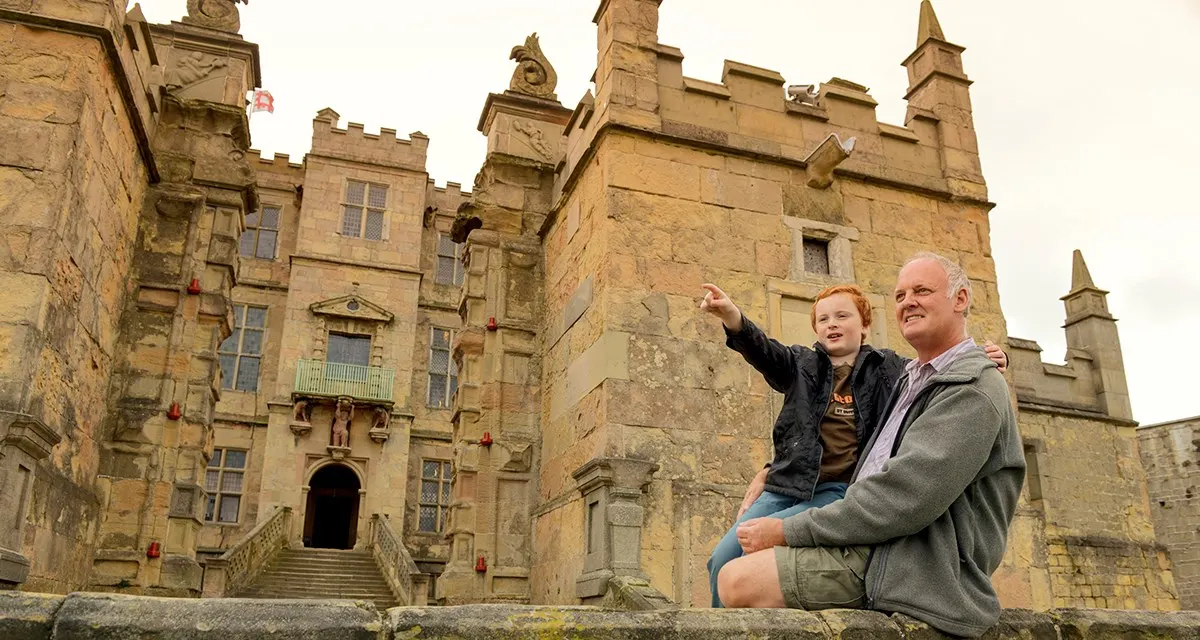 An adult and child sit on a stone wall pointing at a large sandstone castle with multiple turrets and castellated towers.