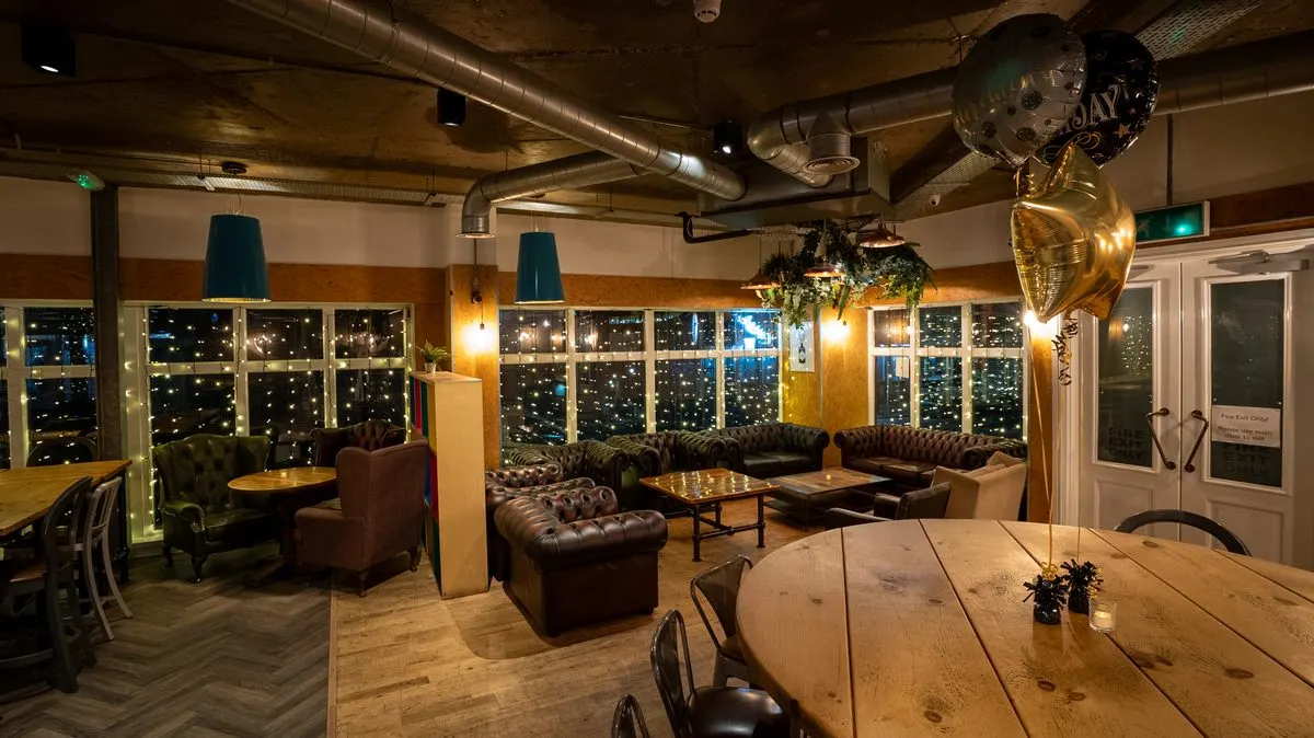 Interior of a modern events venue with wooden tables, leather seating, floor-to-ceiling windows overlooking a night cityscape, and industrial ceiling details.