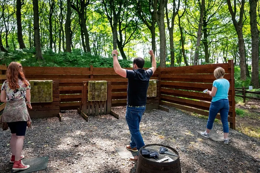 A man stands on a tyre doing an axe throw while two children watch in a woodland clearing with wooden barriers.