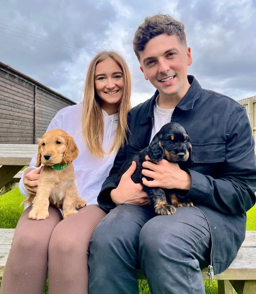 A young man and woman posing outdoors on a wooden deck, each holding a puppy.