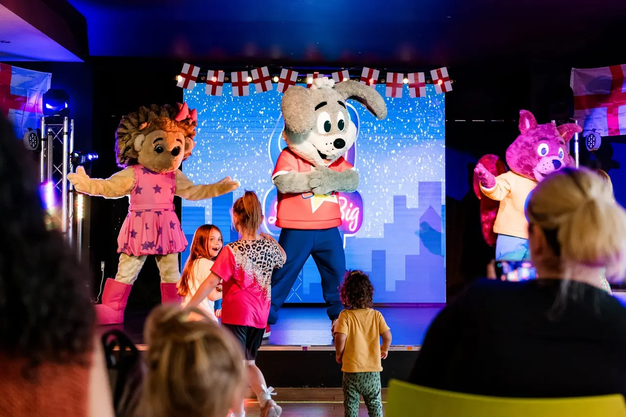 Children watching costumed character mascots perform on an illuminated stage with festive decorations and flags.