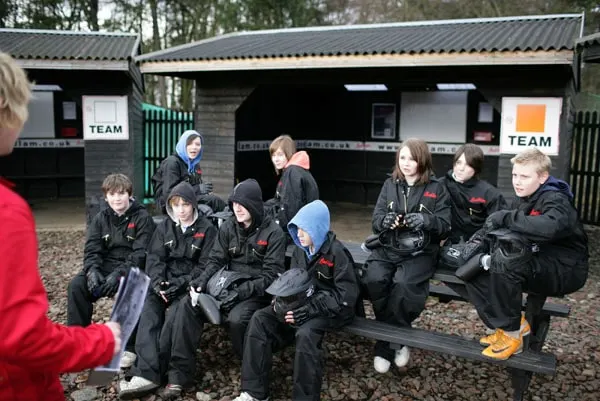 A group of players in black paintball gear sit and stand outside a dark wooden building, preparing for a paintball game.