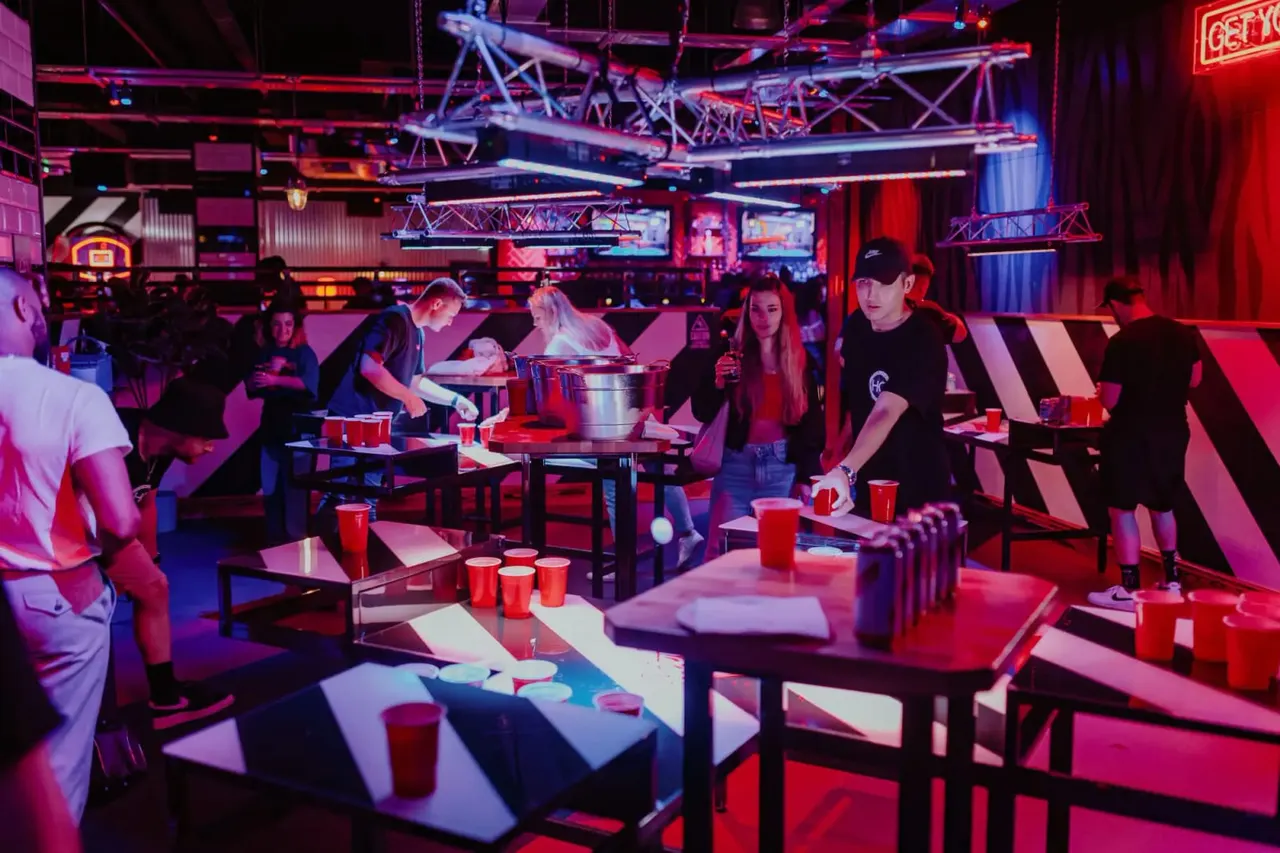 Groups of people playing beer pong and other games in a neon-lit indoor entertainment venue with red cups and tables.