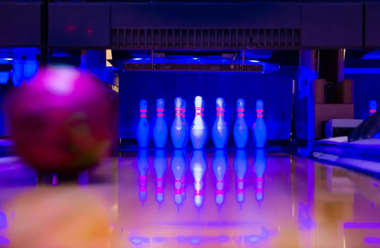 Bowling alley with illuminated pins set up on the lane and a glowing ball in the foreground under blue and pink lighting.