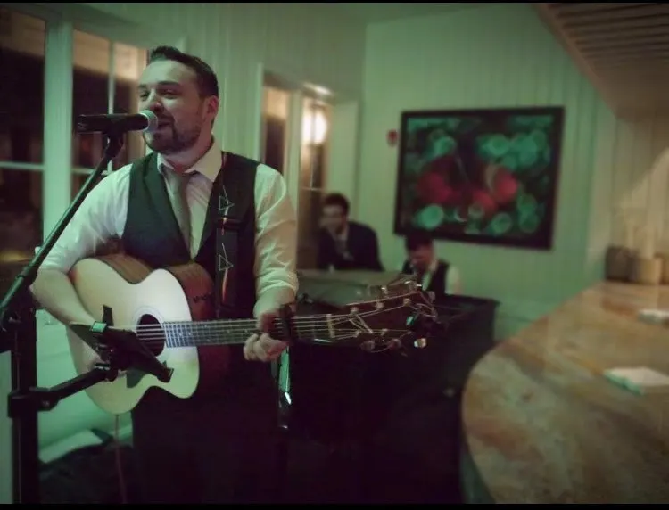 A man plays acoustic guitar and sings at a microphone in an indoor venue with green walls whilst seated guests watch.