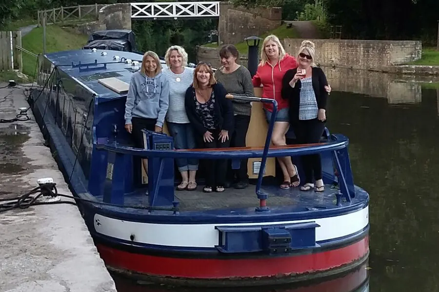 Six women stand aboard a blue and red narrowboat moored on a canal, with countryside and wooden gates visible beyond.