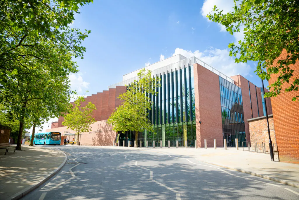Modern brick and glass theatre building with green trees framing the courtyard entrance on a sunny day.