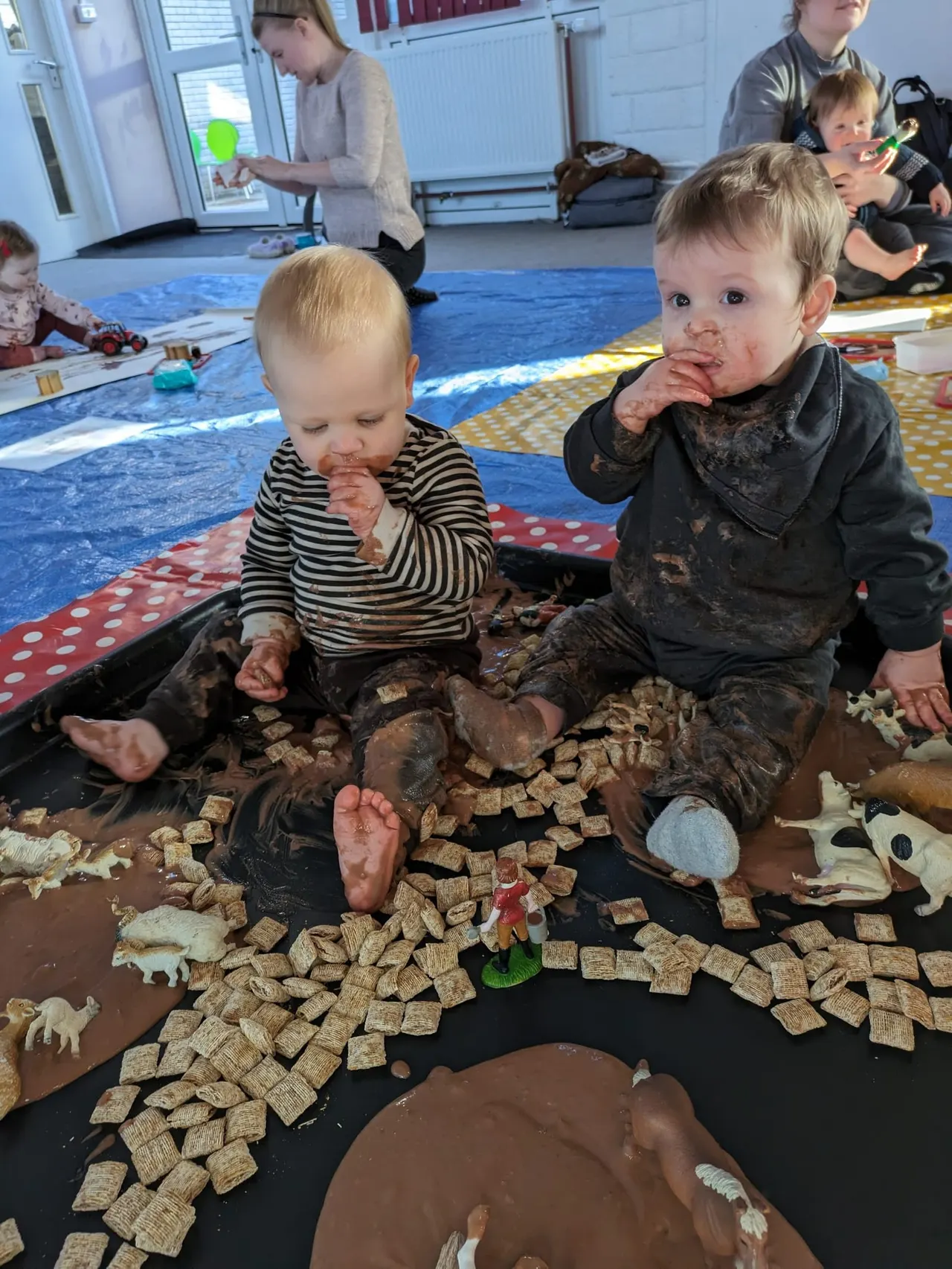 Toddlers play in a sensory mud and cereal activity whilst other children play on a blue mat in an indoor soft play room.