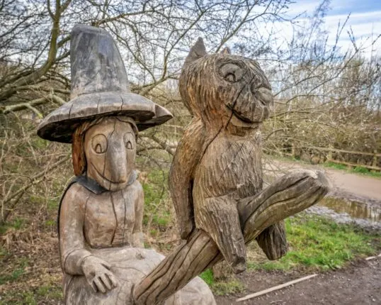 Wooden carved figures of a character wearing a hat and a squirrel displayed outdoors among bare trees.