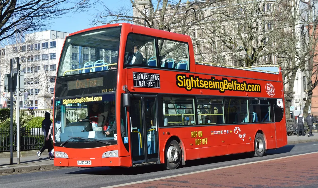 A red double-decker sightseeing bus stopped on a city street with passengers visible through the windows and bare trees in the background.
