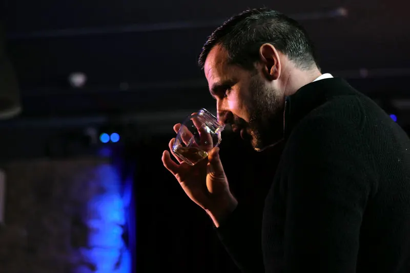 Man in black shirt holding a glass of whisky in a dimly lit bar with blue ambient lighting.
