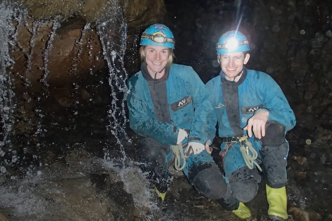 Two people wearing blue helmets and climbing gear smile at the camera inside a cave with water flowing down the rock walls.