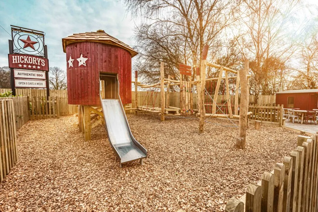Children's play area with red wooden structures, slide, climbing frames and bare trees in winter at a leisure venue.