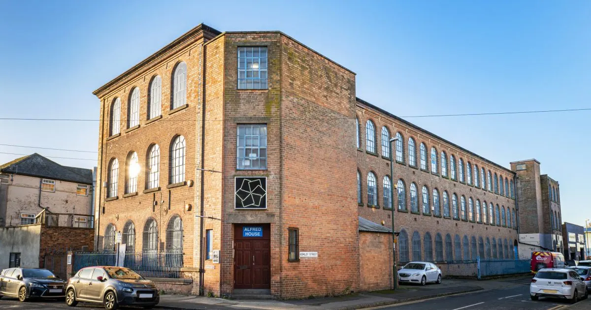 Historic brick building with large arched windows and parked cars on street below, clear blue sky.