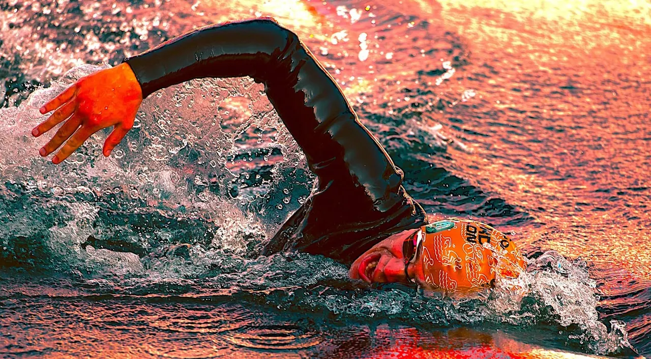 A swimmer in red swimming cap and orange arm floats performs freestyle stroke in sparkling water at sunset.