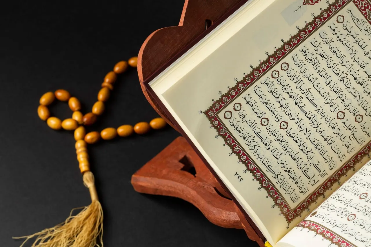 An open Qur'an with decorative border resting on a wooden stand, alongside a beaded prayer cord with tassel.