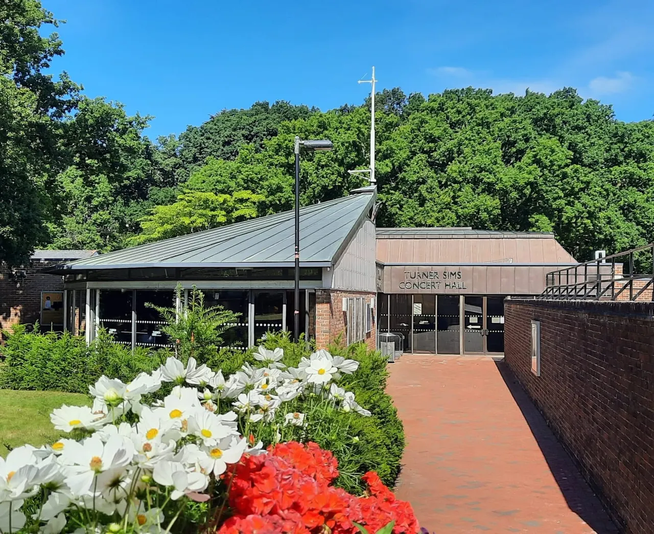 Modern concert hall building with metal roof and cross, surrounded by flowering gardens with white daisies and red blooms, set against woodland backdrop on a sunny day.